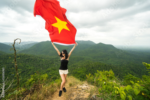 Beautiful young woman is standing on top of the mountain and holding vietnamese flag