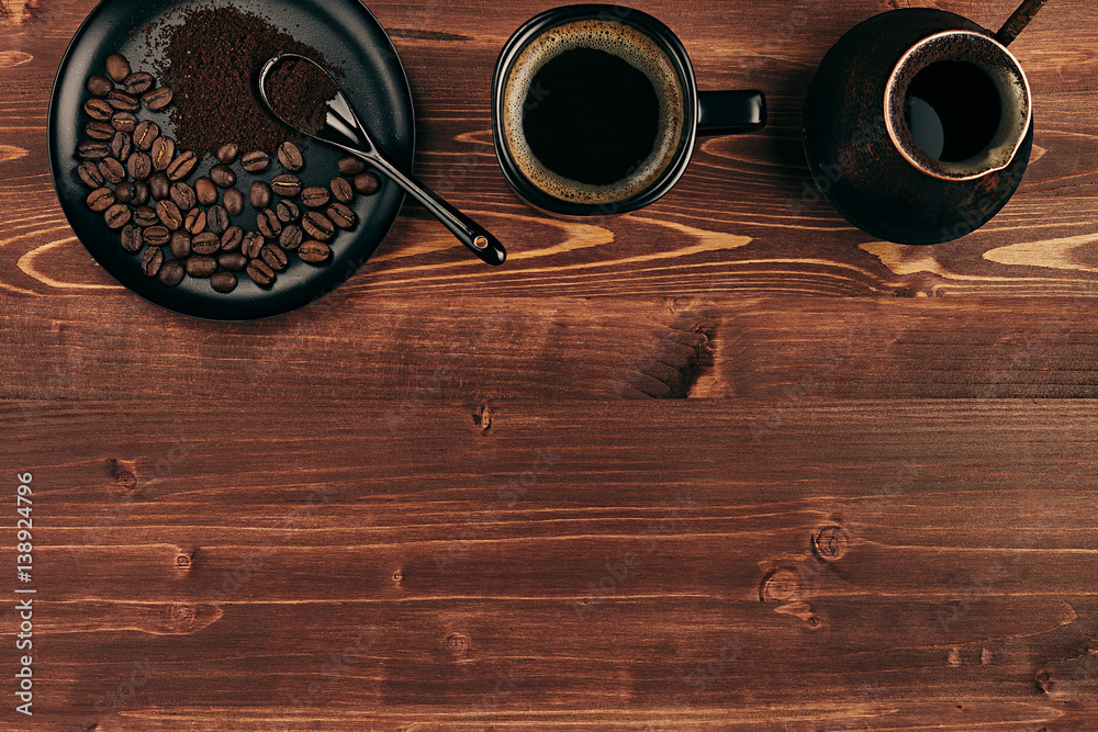Hot coffee in black cup with beans, spoon and turkish pot cezve with copy space on brown old wooden board background, top view. Rustic style. Hot coffee in black cup with beans, spoon and turkish pot cezve with copy space on brown old wooden board background, top view. Rustic style.