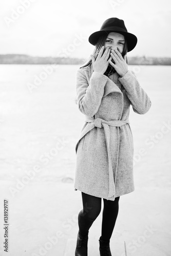 Young model girl in gray coat and black hat  posed against freeze lake. © AS Photo Family
