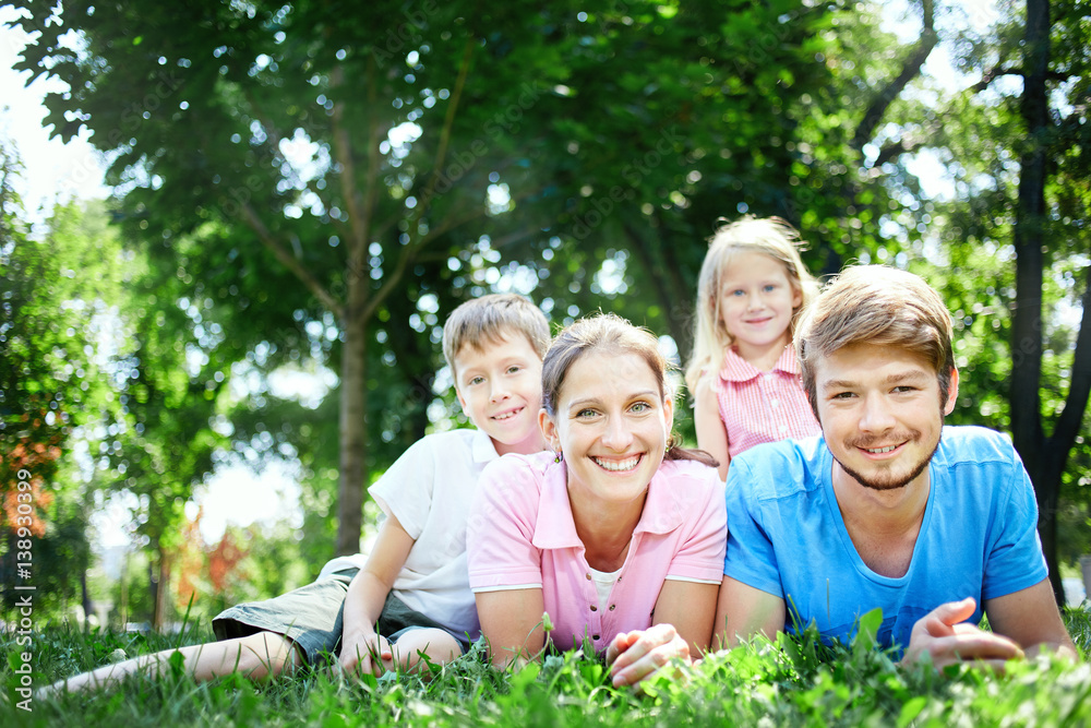 Fototapeta premium young happy family at noon in the park on the grass. Two young parents and children, boy and girl, lies on the grass and smiling looking at the camera.