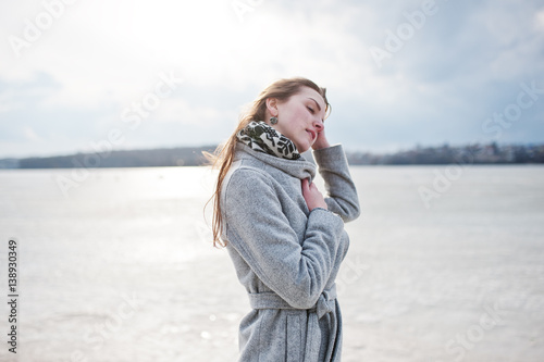 Young model girl in gray coat with red hair against freeze lake.
