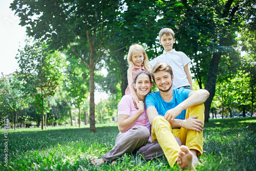 young happy family at noon in the park on the grass. Two young parents and children, boy and girl, sits on the grass and smiling looking at the camera.