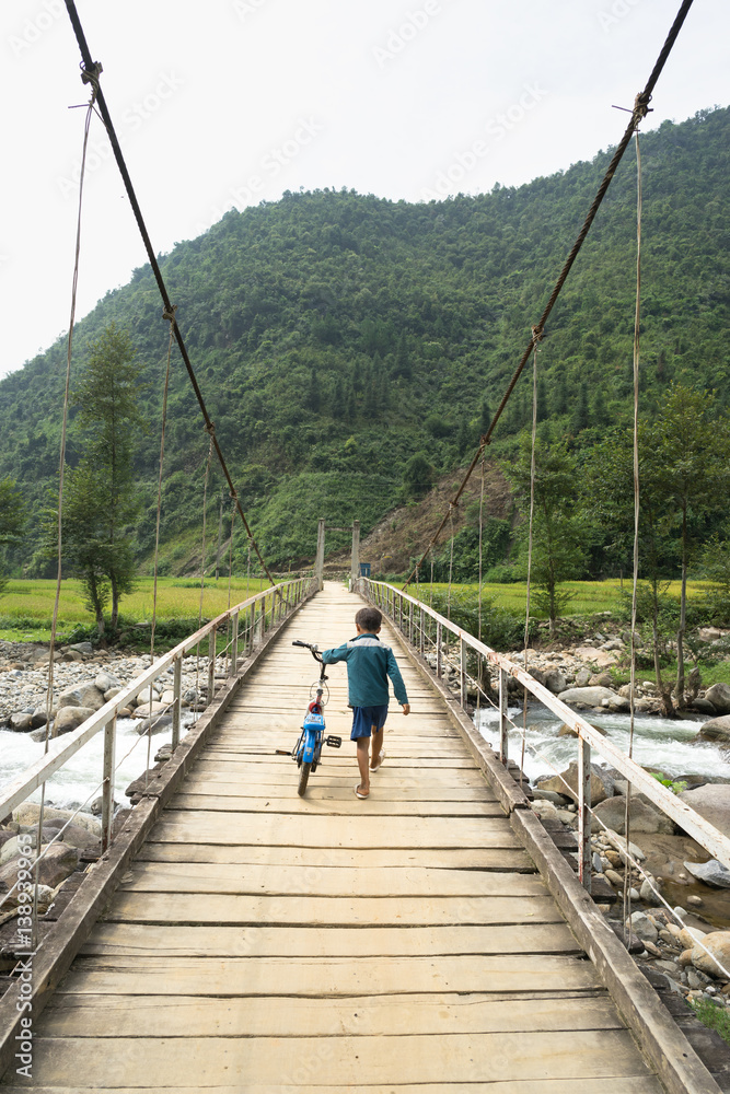Vietnamese Hmong ethnic minority boy walking on old wooden bridge with his bicycle