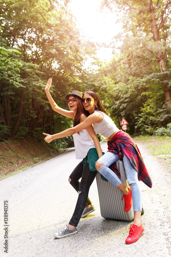 Two young tourist girl hitchhiking by the road.they sitting on suitcase and making fun. © BalanceFormCreative