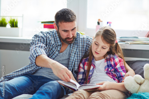 Handsome middle-aged father explaining paragraph presented in textbook to his teenage daughter, she listening to him with interest
