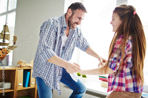Smiling dad and his little daughter taking pleasure in spending weekend together: they holding hands and jumping against window