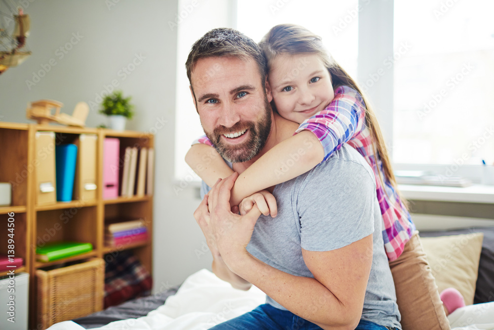 Family portrait in bedroom: little girl in checked shirt leaning on back of her handsome father and looking at camera Family portrait in bedroom: little girl in checked shirt leaning on back of her handsome father and looking at camera