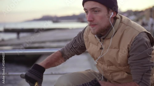 Young Caucasian male with light beard and sock cap putting on headphones sitting on bmx bicycle on pier nea sea then riding away in slowmotion photo
