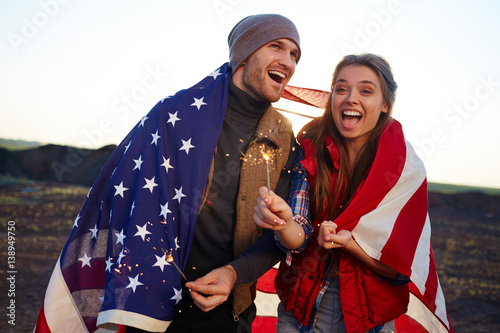 Portrait of young happy couple laughing and holding sparklers, both wrapped in American flag at sunset in mountains