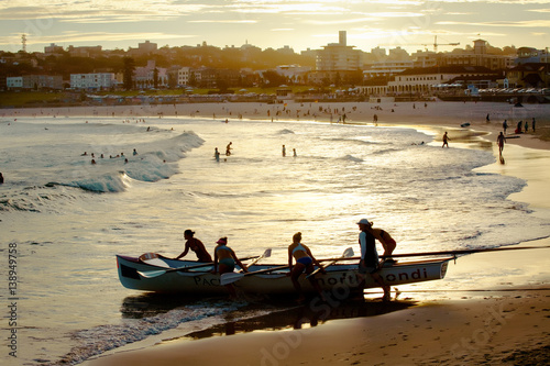 Canvas Print Team of girls push a surfboat in the water at bondi beach sourranding by the sun