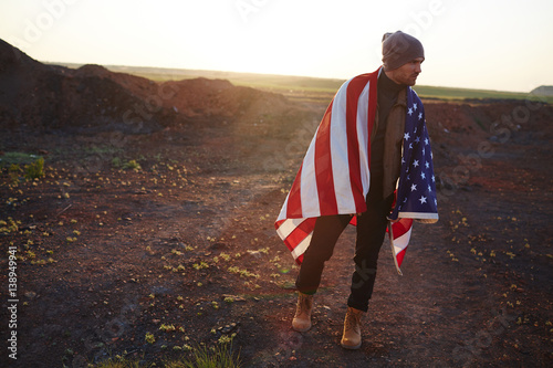Backlit portrait of young fashionable man walking alone at hike in mountains against sunset sky wrapped in big American banner