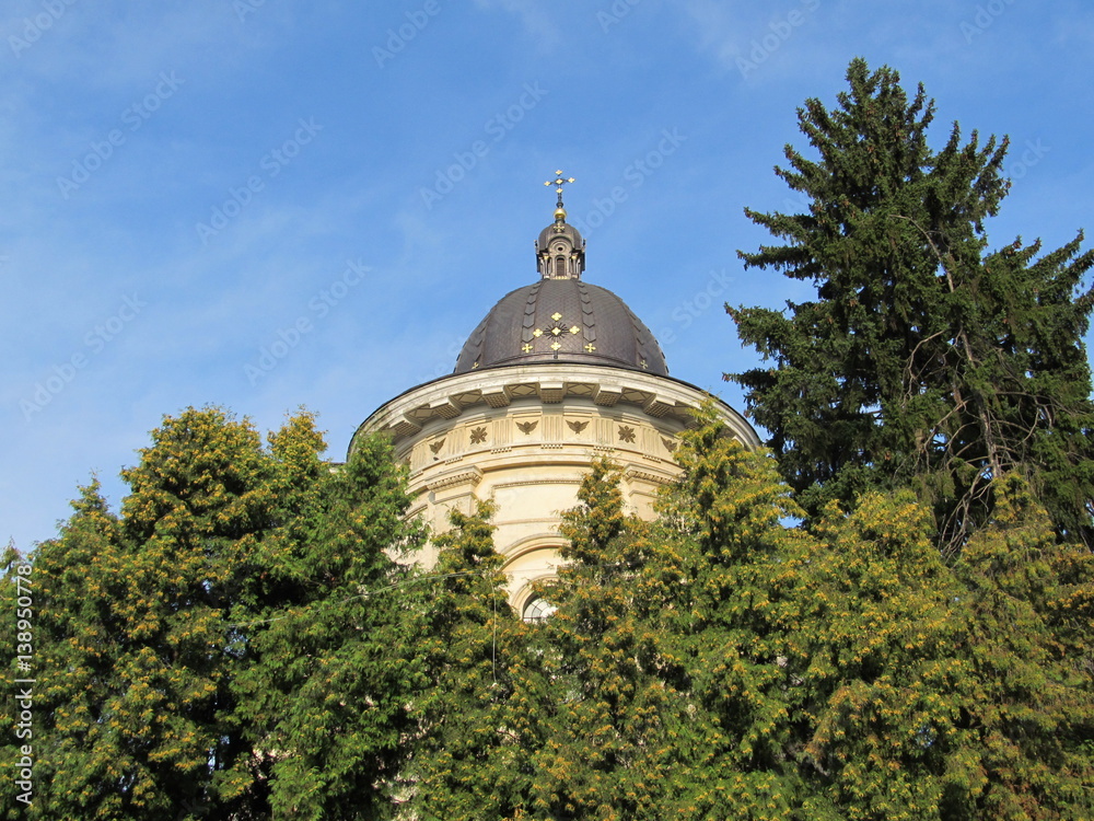 Unusual detail of a top church with cross on the roof against the blue sky in backgrounds and green trees in the foreground