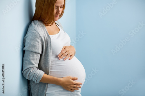 Obraz na plátně Blond pregnant woman leaning on to blue wall caressing her big belly gently