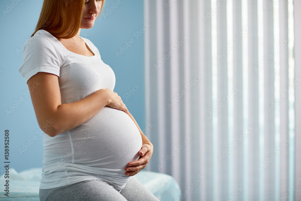 Woman with big belly in her last months of pregnancy sitting on bed in doctors office against window waiting for her examination - obrazy, fototapety, plakaty 