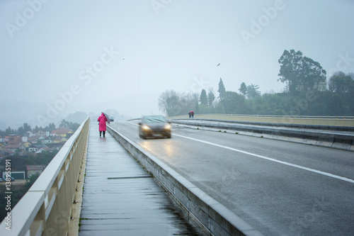 Storm weather. A woman in a pink coat with an umbrella is a sidewalk in the rain. Cars at high speed pass by. © Sodel Vladyslav