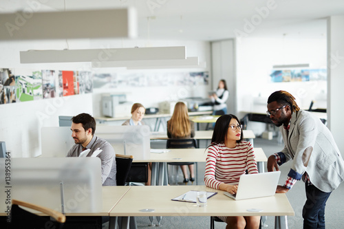 Interior of busy open plan office: male and female workers sitting at their desks, Afro-American superior discussing business project with pretty Asian employee photo