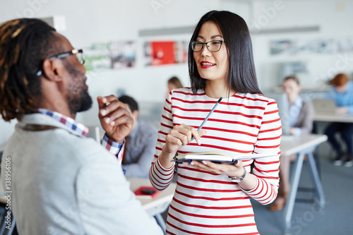 Afro-American boss with short dreadlocks giving instructions to his pretty Asian secretary, she taking notes while standing in open plan office photo