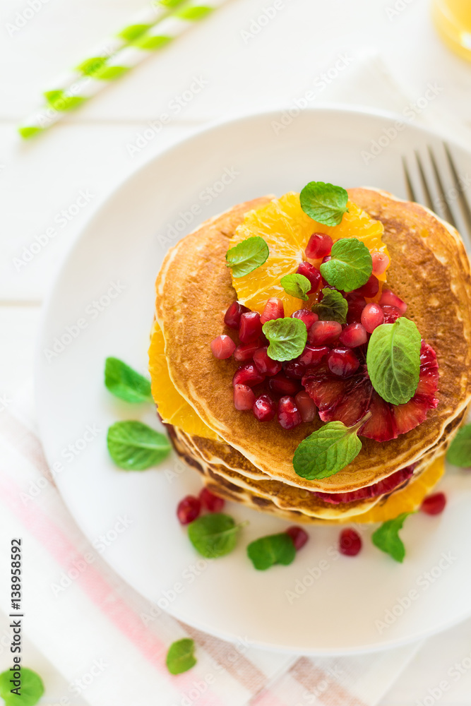 Traditional breakfast: stack of pancakes with orange slices and pomegranate seeds decorated mint leaves on white wooden table. Selective focus Traditional breakfast: stack of pancakes with orange slices and pomegranate seeds decorated mint leaves on white wooden table. Selective focus