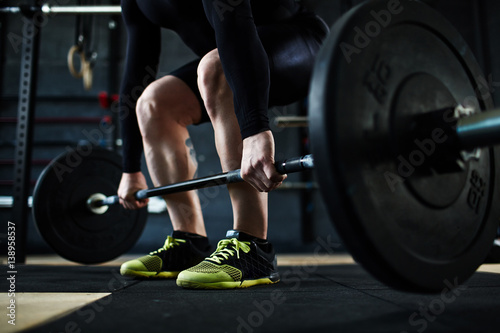 Low section of unrecognizable male athlete lifting huge heavy barbell from floor, leg muscles straining with effort