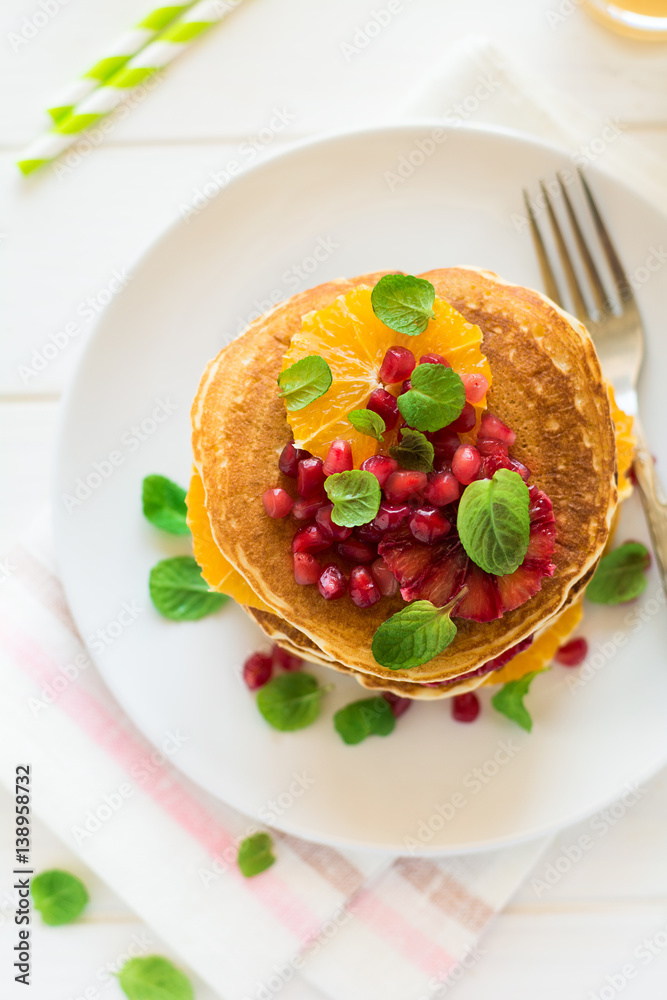 Traditional breakfast: stack of pancakes with orange slices and pomegranate seeds decorated mint leaves on white wooden table. Selective focus Traditional breakfast: stack of pancakes with orange slices and pomegranate seeds decorated mint leaves on white wooden table. Selective focus