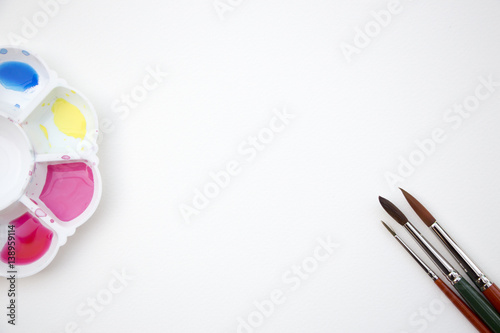 A watercolor dish palette filled with paints and brushes placed on blank white paper as background