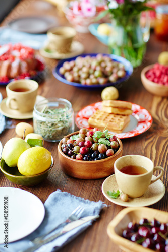Breakfast table setting full of gooseberries and currant, fruits, freshly baked waffles and cups of scented tea