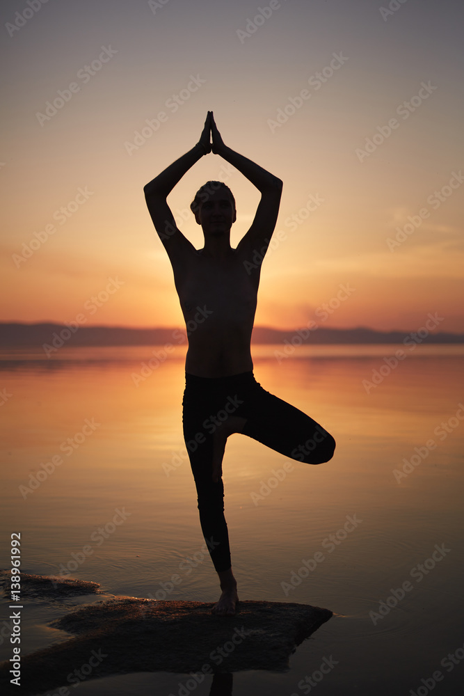 Silhouette of man practicing yoga pose on beach, standing on one leg and holding hands in namaste