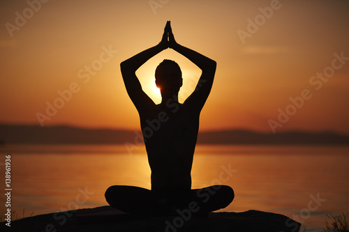 Silhouette of man sitting in lotus pose with his arms lifted above head in namaste and enjoying sunset on water background