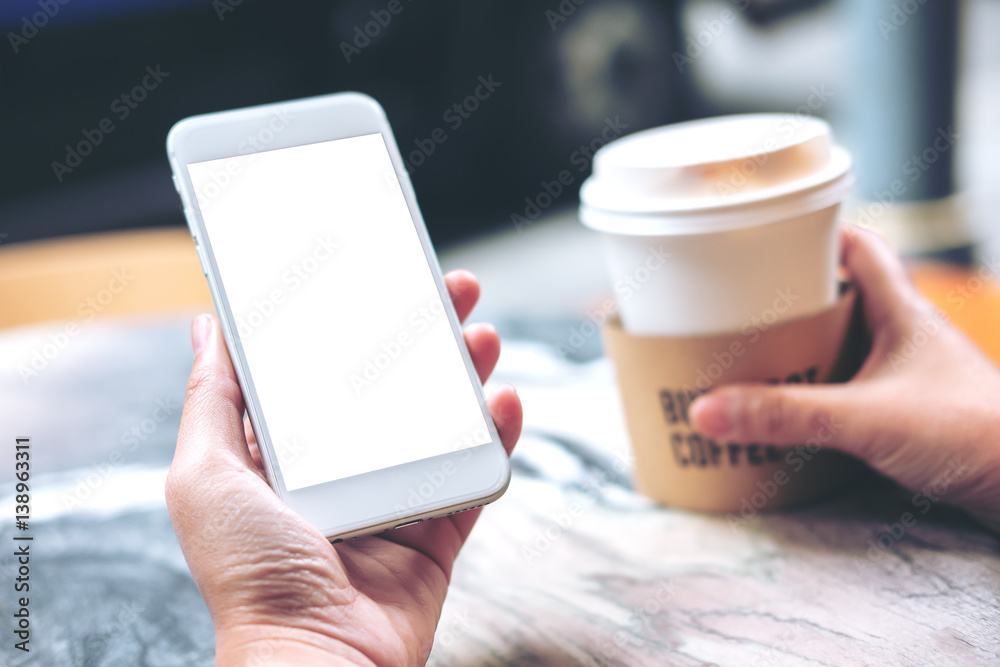 Mockup image of hand holding white mobile phone with blank white screen and coffee cup on marble table in cafe