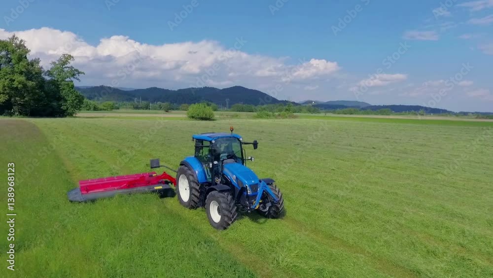 We can see a blue tractor driving on the field and it is cutting grass with red agricultural machinery. It is a nice summer day.
We can see a blue tractor driving on the field and it is cutting grass with red agricultural machinery. It is a nice summer day.
