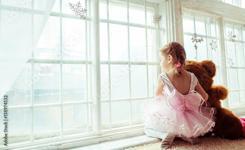 Little girl in pink dress sits with toy bear before panoramic window
