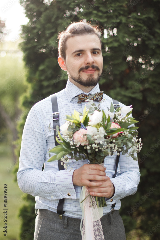 Portrait of attractive young elegant groom dressed wedding tuxedo costume is waiting for the bride with bouquet of white peonies. Wedding photo session of the handsome fiance in a suit at the studio