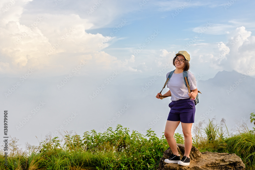 Tourist teens girl hiker with backpack caps and glasses is standing smile and poses happily at high mountain on sky and fog background at scenic point of Phu Chi Fa Forest Park in Chiang Rai, Thailand Tourist teens girl hiker with backpack caps and glasses is standing smile and poses happily at high mountain on sky and fog background at scenic point of Phu Chi Fa Forest Park in Chiang Rai, Thailand