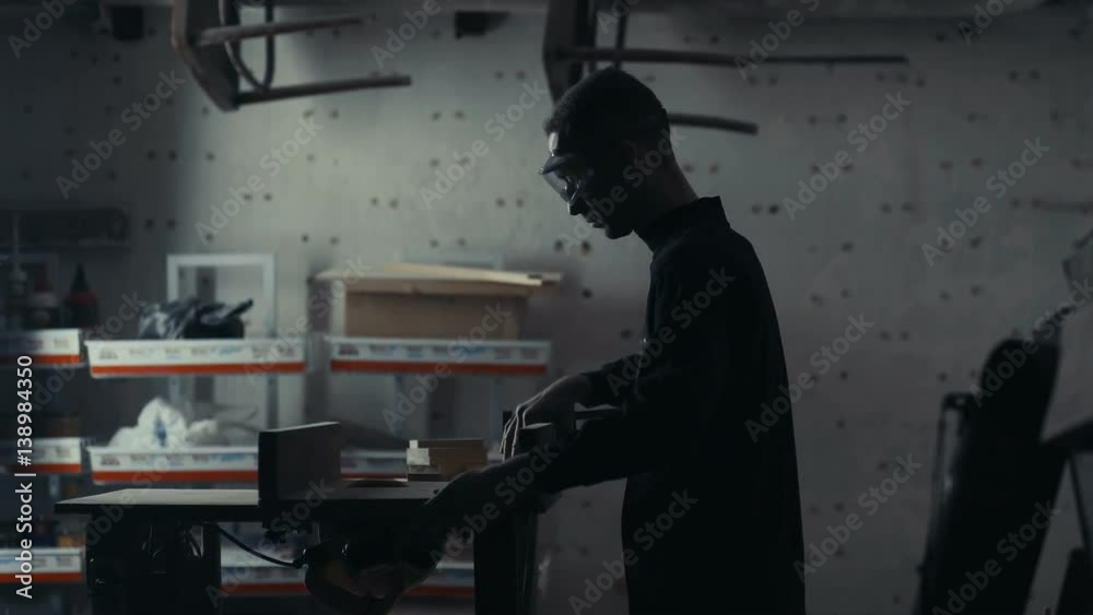Joiner works with an electric jigsaw and processes wooden products. Craftsman with saved glasses work at his workstation. Man in a protective mask on a dark background working on woodworking tools. Joiner works with an electric jigsaw and processes wooden products. Craftsman with saved glasses work at his workstation. Man in a protective mask on a dark background working on woodworking tools.