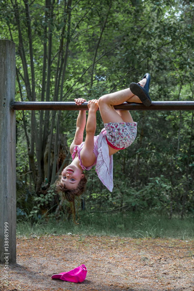 Cute girl in dress and shorts hanging up side down at a climbing frame  outdoors, summer activity. Stock Photo