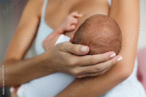 Happy young mother, sitting in the bedroom with her newborn son in her arms,gently supports the child's head with your hand, feeds the baby's chest,the boy greedily sucks her mother's breast photo