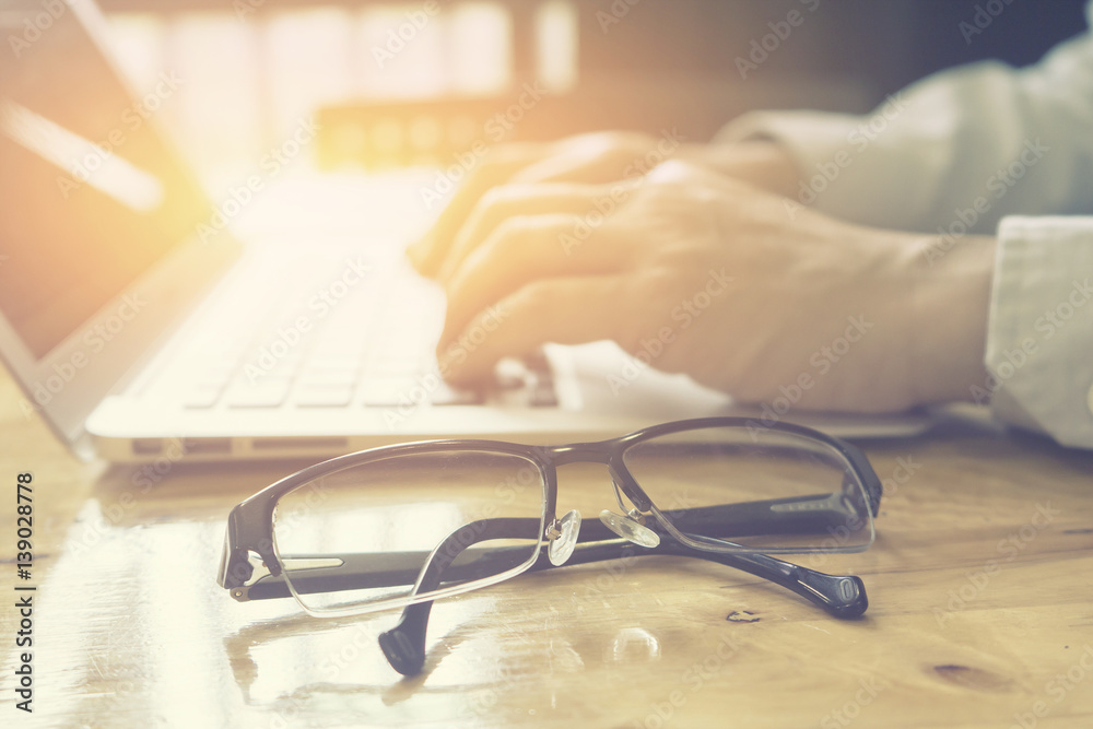 glasses on office desk and office supplies.office desk table with a lot of things on it.and business man typing, writing business plan in background with copy space.vintage color.selective focus... glasses on office desk and office supplies.office desk table with a lot of things on it.and business man typing, writing business plan in background with copy space.vintage color.selective focus...
