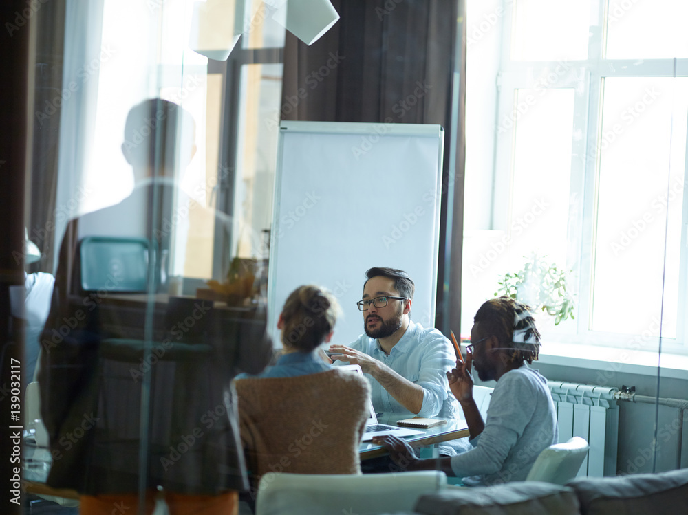 Group of creative young people, Afro-American man among them, sitting round meeting table in modern conference room listening intently to Asian speaker, shot from behind glass wall
