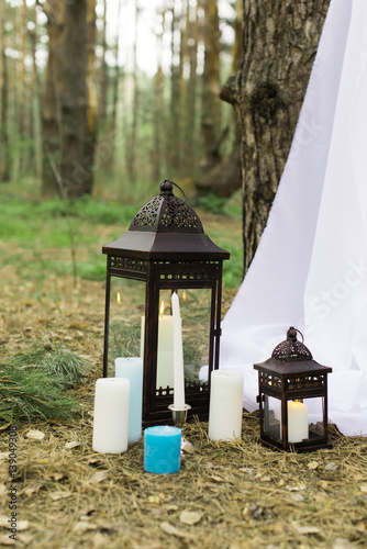 White and blue candles on candlesticks and in lanterns in forest near the wedding arch with white material