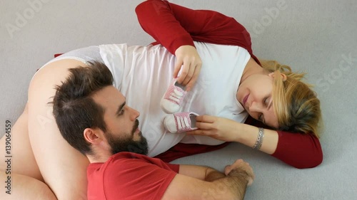 Pregnant woman playing with baby shoes and unconcerned bearded man looking at mobile phone while relaxing on the bed at home.