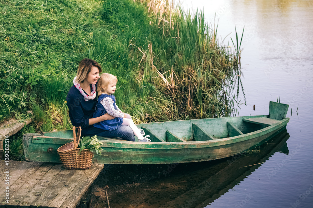 Portrait of happy family of two people on vacation. Young mother and little daughter on picnic sitting in old wooden boat on spring river over water background. Age of child 2 years and 4 month.