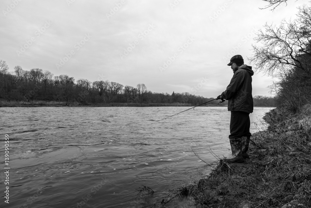 Rugged outdoors fisherman standing on bank of fast flowing river fishing, black and white