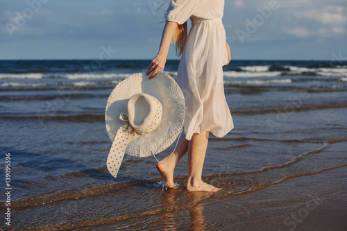 Summer day. Young woman silhouette with big white hat and white dress, legs walking on ocean waves, enjoy of summer travel and nature