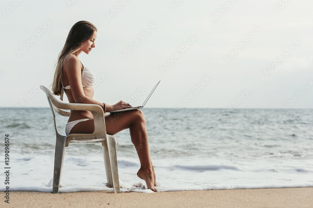 Young woman in white swimsuit using laptop with internet sitting on a chair on tropical island beach. Summer travel, freelance concept, shopping online