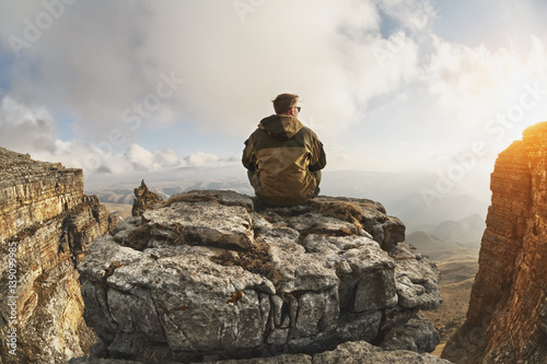 A young man sits on a rock surrounded by cliffs  located above the clouds in a great location  on a background of clouds  valleys  fields  mountains