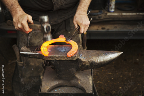 A farrier using tongs and hammer to hold and shape a red glowing heated metal horseshoe to be fitted.