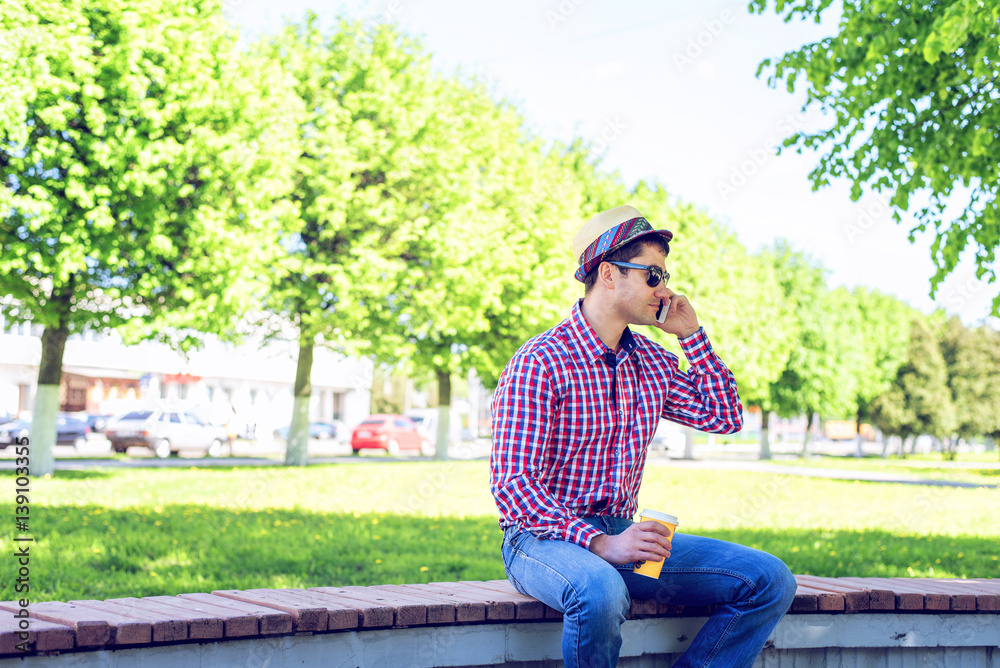 The man is sitting, holding a smart phone, talking on the phone, in the summer in glasses and a shirt with a hat on a bench in the woods outdoors resting businessman The man is sitting, holding a smart phone, talking on the phone, in the summer in glasses and a shirt with a hat on a bench in the woods outdoors resting businessman