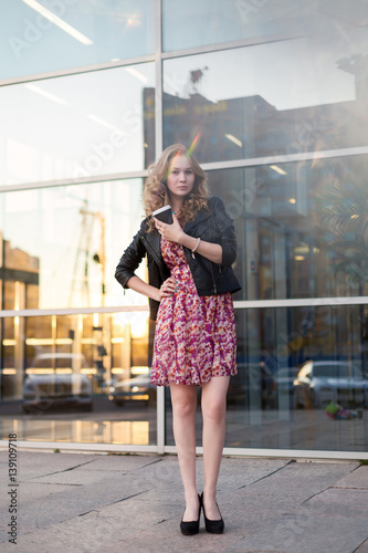 Wallpaper Mural Beautiful blonde girl in dress and black leather jacket stands in the background of the business center with reflective glass windows and drinking coffee from a plastic cup. Torontodigital.ca