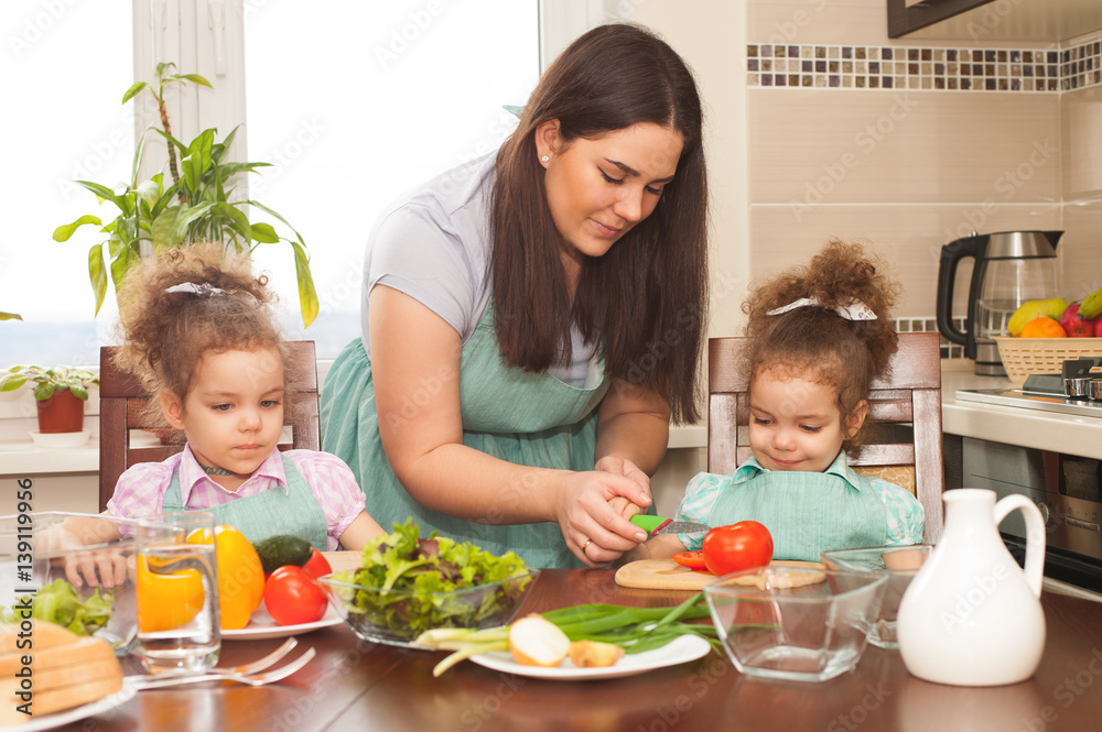 Family preparing meal together. Happy mother and her cute twin daughters having fun cooking vegetable salad.