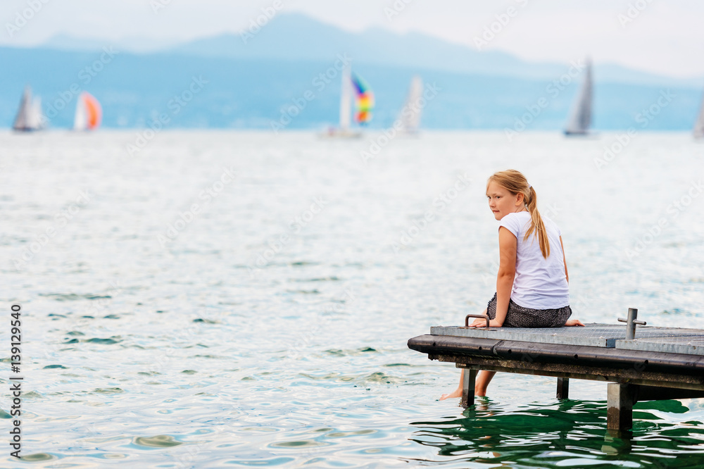 Cute little kid girl resting by the lake, sitting on pier, splashing water with her feet, back view